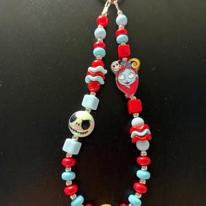 May include: A beaded lanyard featuring a red plastic ring at the top, alternating red and light blue beads, a round white bead with a black Jack Skellington face, a flat charm of Sally, and a single yellow bead with white dots at the bottom.