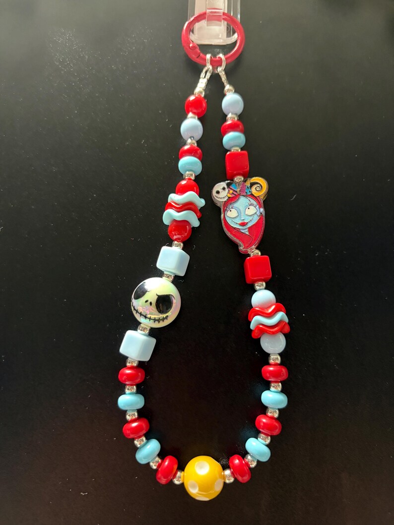 May include: A beaded lanyard featuring a red plastic ring at the top, alternating red and light blue beads, a round white bead with a black Jack Skellington face, a flat charm of Sally, and a single yellow bead with white dots at the bottom.