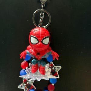 May include: A Spider-Man keychain featuring a small red and blue figurine attached to a silver metal lobster clasp and ring. The figure is surrounded by a circular arrangement of matte red and blue beads, interspersed with two star-shaped charms.