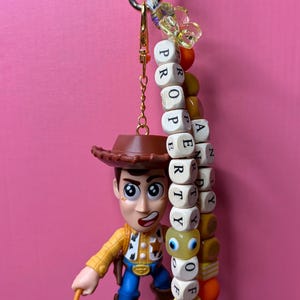 May include: A Woody action figure keyring with a gold-coloured clasp. The keyring has a beaded strand with the word "PROPERTY OF" spelled out in wooden letter blocks. The figure is wearing a cowboy hat, yellow shirt, blue jeans, and brown boots.