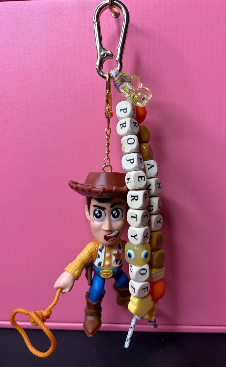 May include: A Woody action figure keyring with a gold-coloured clasp. The keyring has a beaded strand with the word "PROPERTY OF" spelled out in wooden letter blocks. The figure is wearing a cowboy hat, yellow shirt, blue jeans, and brown boots.
