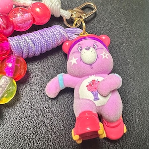 May include: A purple Care Bear plush keychain with a gold clasp, a beaded lanyard, and a purple cord. The Care Bear has a white belly with a dessert graphic, star cheek accents, and red roller skates.