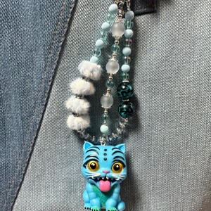 Può includere: Un ciondolo per borsa con una statuetta di tigre azzurra con grandi occhi gialli e lingua rosa, sospesa a una catena di perline a più fili. La catena include perline soffici azzurre, sfere traslucide smerigliate e perline verde acqua scuro con motivi.