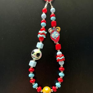 May include: A beaded lanyard featuring a red plastic clip, alternating red and light blue spherical and cube beads, a round bead with a Jack Skellington face, a flat charm of Sally, and a single yellow bead with white polka dots at the bottom.