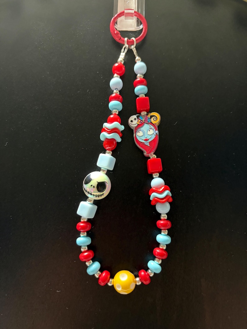 May include: A beaded lanyard featuring a red plastic clip, alternating red and light blue spherical and cube beads, a round bead with a Jack Skellington face, a flat charm of Sally, and a single yellow bead with white polka dots at the bottom.