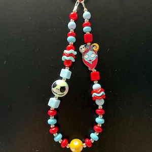 May include: A beaded lanyard featuring a red circular clasp at the top. The strand includes red, light blue, and yellow beads in various shapes, including round, square, and wavy designs. It features a character charm of Sally and Jack Skellington.