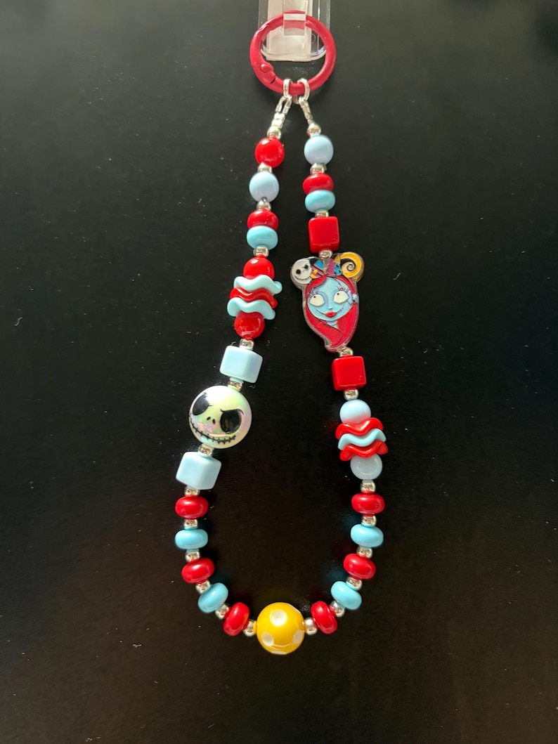 May include: A beaded lanyard featuring a red circular clasp at the top. The strand includes red, light blue, and yellow beads in various shapes, including round, square, and wavy designs. It features a character charm of Sally and Jack Skellington.