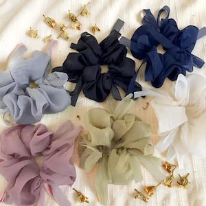 May include: Six large scrunchies in a range of colours, including navy, black, white, lavender, and sage green. Each scrunchie is made from sheer fabric and has long, flowing ribbons. Dried flowers are scattered around the scrunchies.