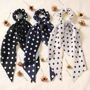 Peut inclure: Trois chouchous à pois avec de longues queues flottantes. L'un est noir à pois blancs, un autre est bleu marine à pois blancs, et le troisième est blanc à pois noirs. Ils sont disposés sur une surface claire avec des fleurs séchées.