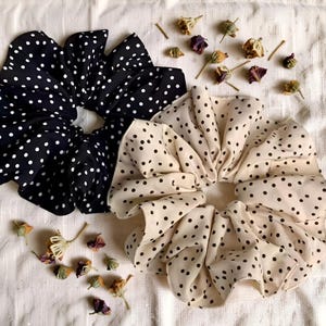 Peut inclure: Deux grands chouchous, l'un noir à pois blancs et l'autre crème à pois noirs, sont présentés sur une surface blanche. Des fleurs séchées sont dispersées autour des chouchous, ajoutant une touche de décoration naturelle.