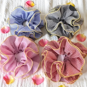 Könnte beinhalten: Vier gerüschte Haargummis in blauem, schwarzem, rosa und rotem Vichy-Muster mit gelbem Besatz. Die Haargummis liegen auf einer weißen Oberfläche, umgeben von verstreuten rosa und gelben Blütenblättern.