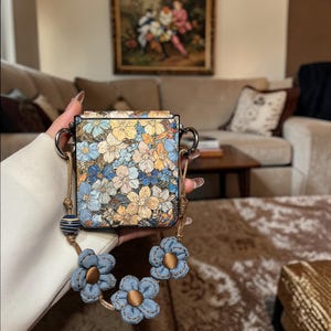 May include: A square handbag with a floral pattern in shades of blue, yellow, and cream. The bag has silver-toned handles and a detachable strap with denim flower accents. The strap includes a decorative blue and white striped bead.