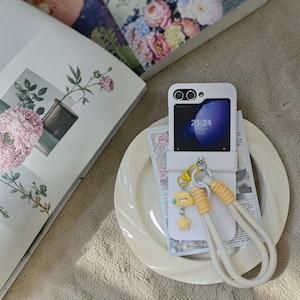 May include: White phone case with a screen displaying 21:24. The case has a yellow and white wrist strap and a charm with a yellow cloud and star. The phone rests on a white plate with a newspaper clipping.