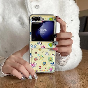 May include: A yellow phone case with a cartoon design featuring various characters and the text "Tamagotchi." The case is open, revealing a phone screen displaying the time. The case has a silver frame.