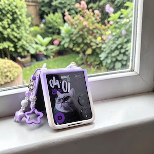 May include: A lilac and white flip phone case with a matching charm. The phone screen shows a cat wearing glasses. The charm includes beads and a star-shaped pendant. The phone is open, displaying the date and time.