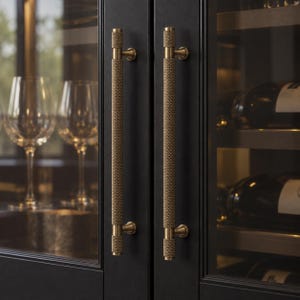 May include: Two vertical brass cabinet handles with a textured, knurled grip pattern. The handles are mounted on a black cabinet door frame, featuring a cylindrical shape with small circular attachment points at the top and bottom of each handle.
