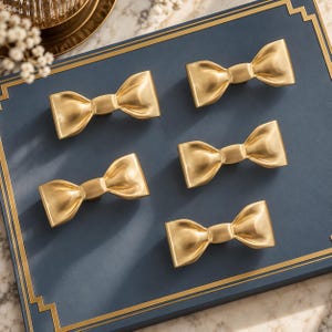 Peut inclure: Un ensemble de cinq boutons de placard en métal doré, chacun en forme de nœud papillon avec une finition lisse et polie. Ces éléments de quincaillerie décoratifs présentent un nœud central et deux extrémités évasées pour les meubles.