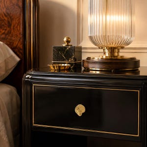 Peut inclure: Une table de chevet noire avec des bordures dorées et une poignée de tiroir en forme de coquillage doré. Sur le dessus se trouvent une lampe en verre strié avec une base en bois sombre, une boîte en marbre noir et un petit plat doré festonné.