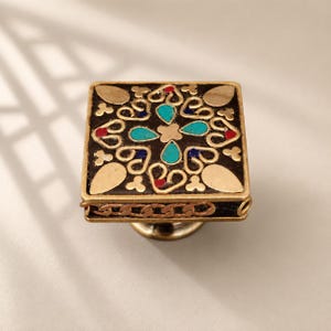 May include: A square cabinet knob featuring a black base with gold-toned metal filigree, inlaid with turquoise, blue, and red accents. The side of the knob is decorated with a metallic chain-link pattern, and it has a circular mounting base.