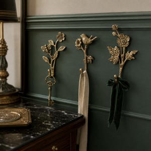May include: Three antique brass wall hooks featuring botanical designs. The left hook depicts a bee and flower, the center hook features a bird on a branch, and the right hook shows a floral spray. Each hook has a curved base for hanging items.