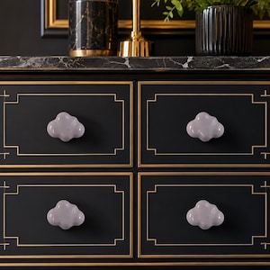 May include: Four light gray, cloud-shaped drawer knobs are mounted on a black dresser with gold trim. Each knob features a smooth, rounded surface and a distinct puffy silhouette, providing a functional handle for the four rectangular drawers.