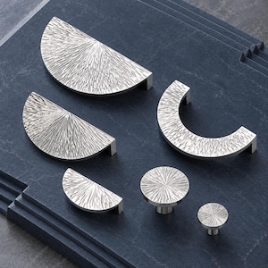 May include: A collection of six silver-toned metal cabinet hardware pieces featuring a textured, sunburst-like pattern. The set includes two large semi-circular pulls, one C-shaped pull, one small semi-circular pull, and two round knobs.