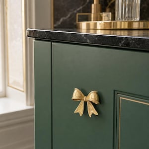 May include: A gold-toned metal cabinet knob shaped like a decorative bow with a textured, beaded edge detail. The bow is mounted on a solid, matte dark green cabinet door surface, featuring a smooth finish and a classic, symmetrical design.