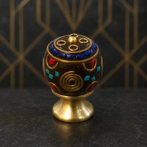 May include: A round cabinet knob featuring a dark brown base with inlaid blue, red, and turquoise stone accents. The knob is decorated with gold-toned metal spiral patterns and circular rings, set upon a polished brass-coloured pedestal base.