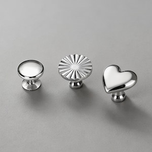 May include: Three polished silver cabinet knobs. One is a clear, round knob, the second has a fluted design, and the third is heart-shaped. All knobs have a short, cylindrical base and are set against a grey background.