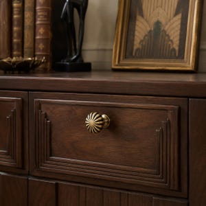 May include: A dark brown wooden drawer featuring a round, fluted brass knob with a sunburst pattern. The drawer front is framed by stepped, rectangular moulding details, showcasing a smooth, polished wood grain finish on the cabinet surface.