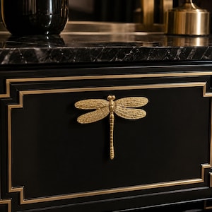 May include: A gold-colored dragonfly-shaped cabinet pull mounted on a matte black drawer front. The dragonfly features a textured wing pattern and a slender, elongated body, centered on the rectangular drawer panel with gold-toned trim.