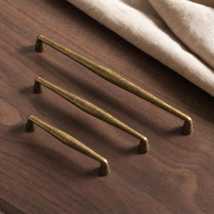 May include: Three metal cabinet pulls in a graduated antique brass finish. Each handle features a slender, slightly tapered cylindrical bar with a textured surface and two small conical mounting posts at the ends, arranged in a diagonal row.