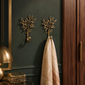May include: Two gold-toned metal wall hooks featuring a decorative branch design with leaves and flowers. One hook is empty, while the other holds a light-colored fabric scarf with a subtle pattern. The hooks are mounted on a dark green wall.