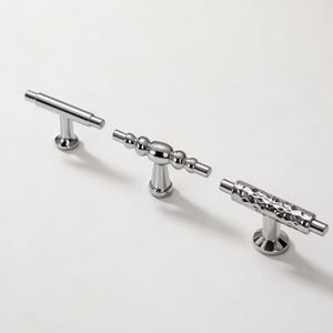 Könnte beinhalten: Drei polierte Chrom-Schrankgriffe in verschiedenen Designs. Einer ist ein einfacher Stab, ein anderer hat eine Reihe von Kugeln und der dritte hat eine strukturierte Oberfläche. Alle haben ein T-förmiges Design und sind auf einem runden Sockel montiert.