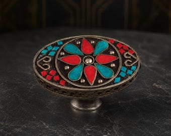 May include: An oval-shaped metal cabinet knob featuring a central floral motif with alternating red and turquoise teardrop-shaped inlays. The surrounding border is decorated with small red and turquoise square tiles and silver heart-shaped accents.