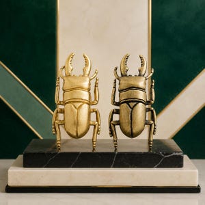 May include: Two metallic beetle figurines with prominent mandibles, one in a bright gold finish and the other in an antique brass finish. Both insects are positioned upright on a rectangular black marble base with white veining.