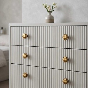 May include: A light grey three-drawer chest of drawers with vertical ribbed detailing. The chest has four gold-coloured knobs and a small vase with white flowers on top. The chest is a modern style.
