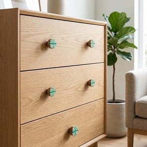 May include: A light-colored wooden dresser with four drawers. Each drawer has a round knob with a green cross design. A potted plant is visible in the background.
