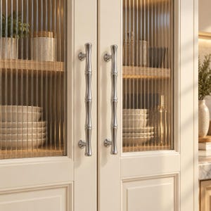 May include: Two polished silver cabinet handles with a segmented bamboo-like design mounted on off-white cabinet doors featuring vertical reeded glass panels that partially obscure the interior shelves containing stacked ceramic bowls and jars.