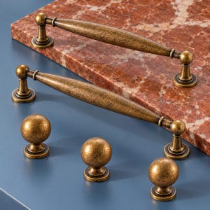 May include: Two elongated, tapered cabinet pulls and three spherical cabinet knobs, all featuring a distressed, antique brass finish with a textured, weathered appearance. The hardware components have circular bases and a consistent metallic gold-brown tone.