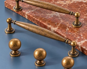 May include: Two elongated, tapered cabinet pulls and three spherical cabinet knobs, all featuring a distressed, antique brass finish with a textured, weathered appearance. The hardware components have circular bases and a consistent metallic gold-brown tone.