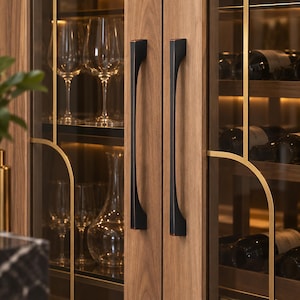 Könnte beinhalten: Ein Weinschrank aus Holz mit zwei vertikalen Griffen aus mattschwarzem Metall. Der Schrank hat Glastüren mit dünnen goldfarbenen Metallrahmen, die Einlegeböden mit Weingläsern, einer Glaskaraffe und Weinflaschen im Inneren zeigen.