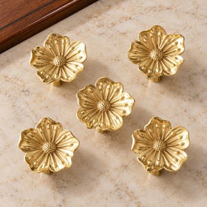 May include: A set of five gold-colored metal cabinet knobs, each shaped like a five-petaled flower with a textured, dotted center and radiating petal details. The knobs have a polished, reflective finish and a uniform, symmetrical design.
