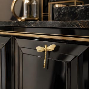 May include: A gold-colored dragonfly-shaped cabinet knob mounted on a glossy black cabinet door. The knob features detailed wings and a segmented body, providing a metallic contrast against the dark, reflective surface of the furniture panel.