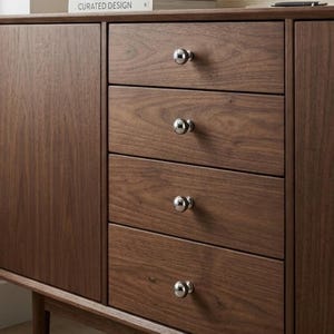 Puede incluir: Un mueble de madera marrón oscuro con cuatro cajones y una puerta lateral. Los cajones tienen pomos redondos y plateados. El mueble es de madera y tiene un diseño moderno. El texto "CURATED DESIGN" es visible en un libro encima del mueble.