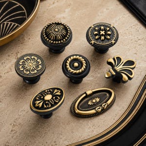 May include: A collection of seven cabinet knobs and pulls in black and antique gold finishes. The hardware features various circular, oval, and fleur-de-lis shapes with intricate embossed patterns, including floral motifs, beaded edges, and rope textures.