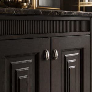 May include: A dark brown wooden cabinet featuring two doors with recessed panels and vertical fluted detailing along the top edge. The cabinet doors are equipped with two polished silver, crescent-shaped metal handles centered on each door.