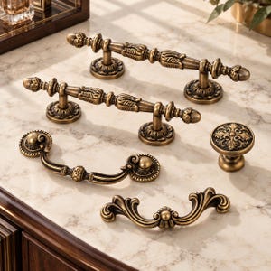 May include: A collection of five antique brass cabinet hardware pieces, including two long straight bar pulls with ornate detailing, one round knob with a floral center, and two curved drawer pulls with decorative scrollwork ends.