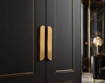 May include: A pair of vertical, matte gold-toned metal cabinet handles with rounded top edges, mounted on double matte black cabinet doors featuring thin, recessed gold-toned trim lines that frame the rectangular panels of the doors.