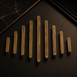 May include: A collection of nine linear cabinet pulls arranged in a symmetrical arc. Each handle features a brushed antique brass finish with rounded ends and a smooth, elongated rectangular profile, varying in length from shortest to longest.